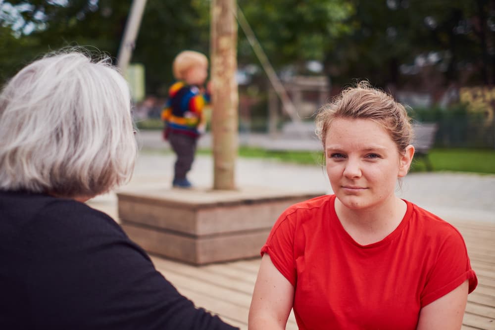 Marit Schatzmann fragend gegenüber einer grauhaarigen Person, im Hintergrund unscharf ein Kleinkind
